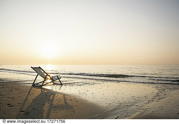 Empty folding chair on shore at beach against clear sky during sunset