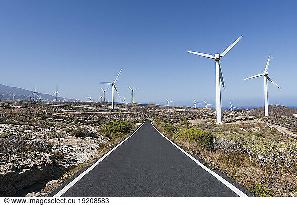 Empty road amidst wind turbines under blue sky