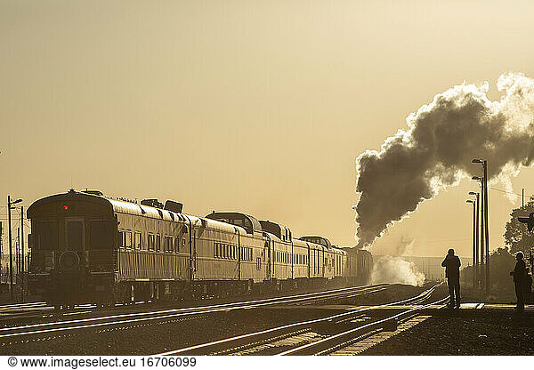 End of a passenger train led by steam engine passes by into yellow sky