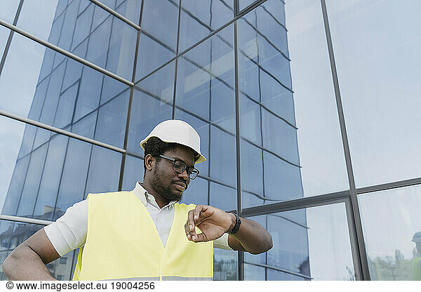Engineer in reflective clothing checking time on wristwatch in front of glass building