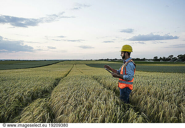 Engineer in reflective clothing holding tablet PC amidst crop in field