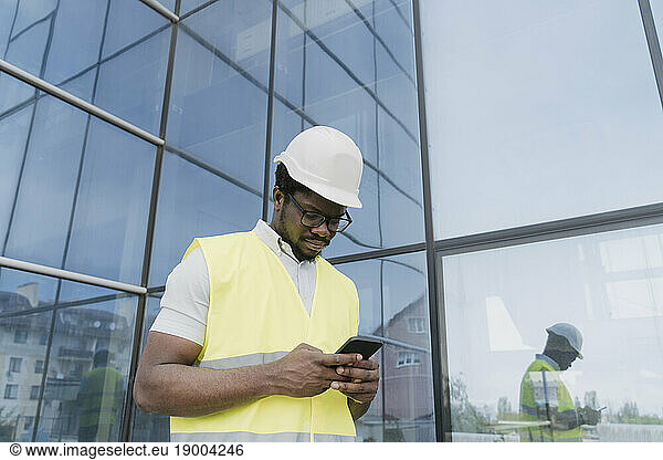 Engineer in reflective clothing text messaging on smart phone in front of glass building