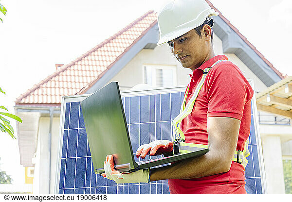 Engineer wearing hardhat using laptop by solar panel in front of house