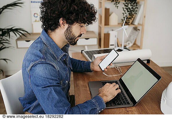 Engineer working on smart phone and laptop sitting at desk in office
