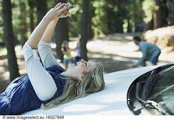 Enthusiastische Frau beim Selbstporträt auf der Motorhaube eines Autos im Wald