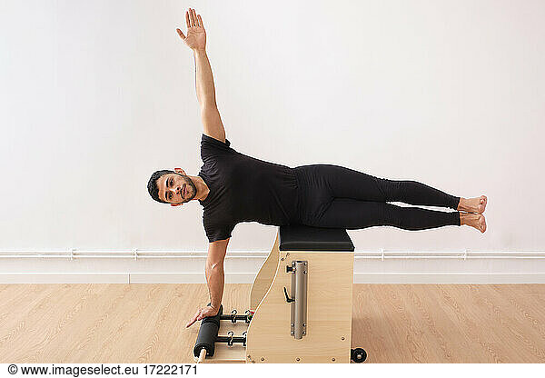 Entschlossener Mann liegt seitwärts mit erhobenem Arm auf einem Pilates-Stuhl im Fitnessraum
