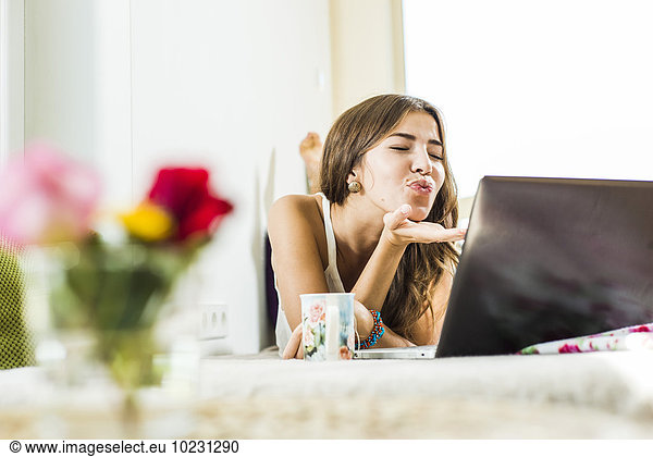 Entspannte junge Frau im Bett liegend mit einem Laptop  der einen Kuss bläst.