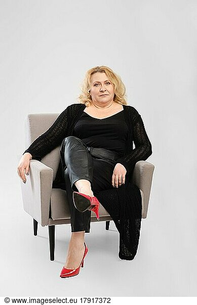 Ernste ältere Frau in Strickjacke und Kunstlederhose sitzt im Sessel vor grauem Studiohintergrund