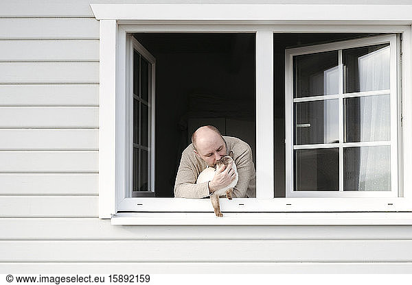 Erwachsener Mann mit Katze  die sich aus dem Fenster seines Hauses lehnt