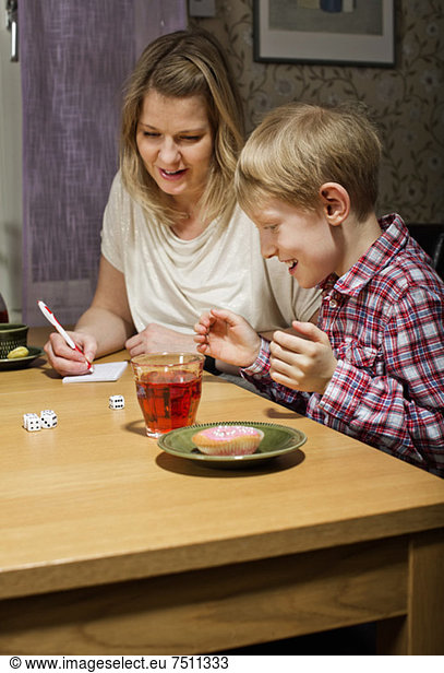 Excited boy playing dice game while mother writing scores at table