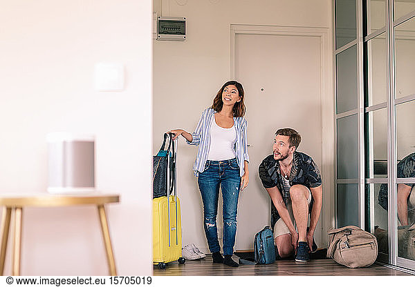 Excited couple with luggage removing shoes while looking around in apartment against doorway