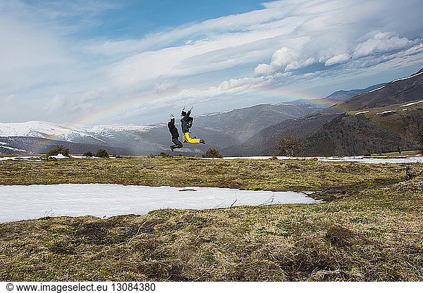 Excited friends jumping against mountains and rainbow during winter