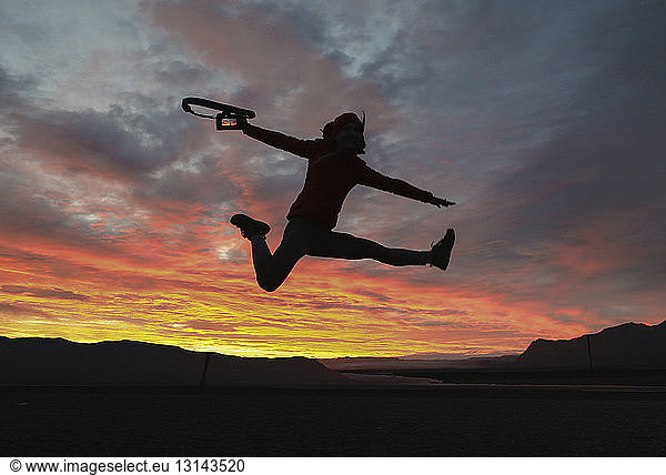 Excited hiker holding camera while jumping against dramatic sky