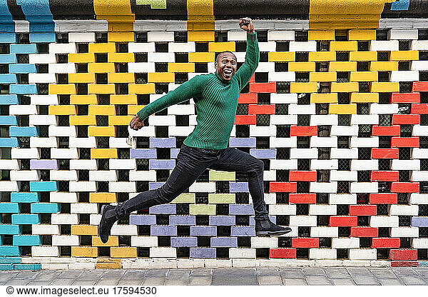 Excited man with hand raised jumping in front of multi colored wall