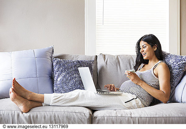 Excited pregnant woman holding bowl while using laptop on sofa