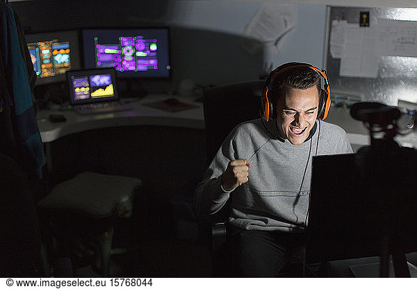 Excited teenage boy with headset cheering  playing video game in dark room