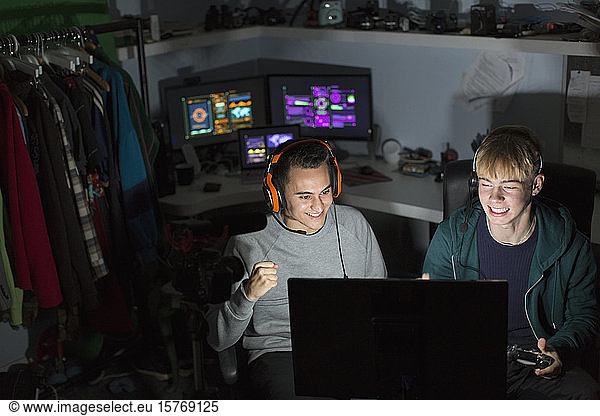 Excited teenage boys with headphones playing video game at computer in dark room