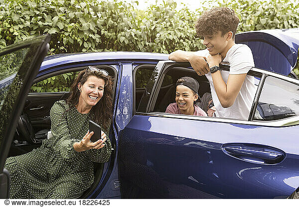 Excited woman sharing smart phone with sons sitting in car