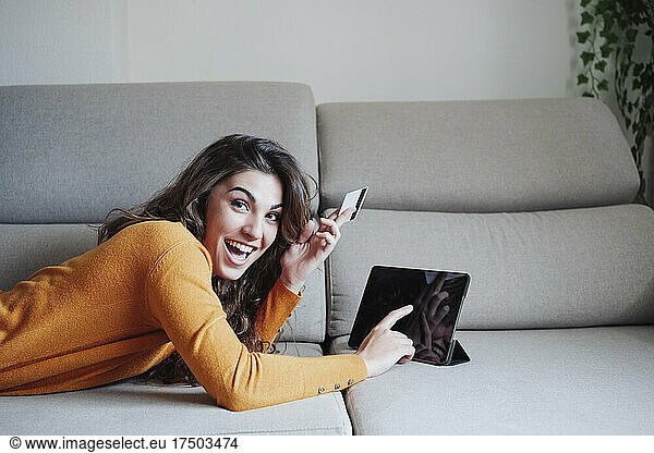 Excited woman using tablet PC holding credit card on sofa at home