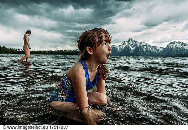 Excited young girl sitting in Jackson Lake with snowy mountains behind