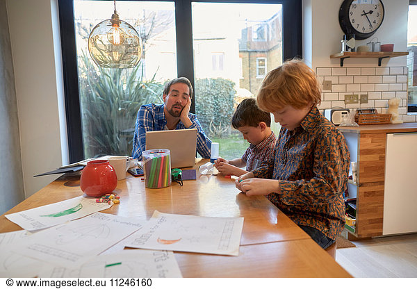 Exhausted man at dining table with laptop whilst son colouring