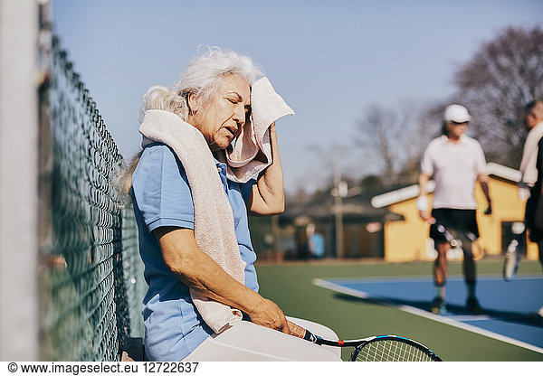 Exhausted senior woman wiping face with towel while sitting at tennis court