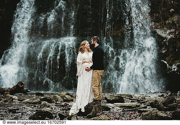 Expecting couple outside in winter by a waterfall standing on rocks