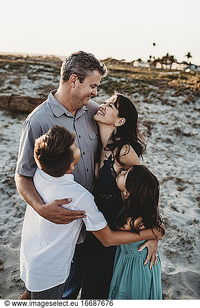 Family hug on sand dune with smiling mid-40's parents and 2 children