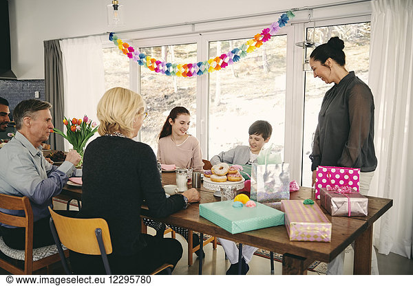 Family looking at boy with gift box while enjoying meal at table during birthday party