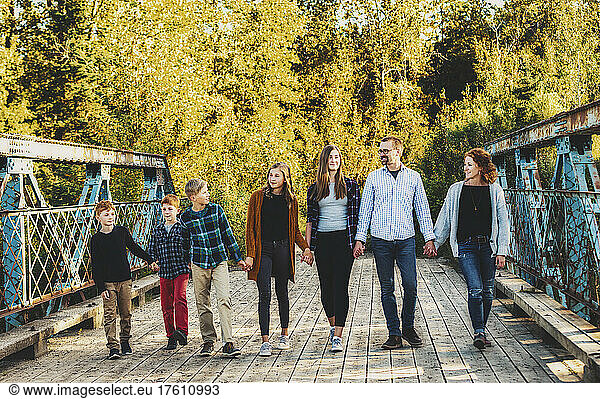 Family of seven walking in a row and holding hands as they walk across a bridge in a city park in autumn; Edmonton  Alberta  Canada