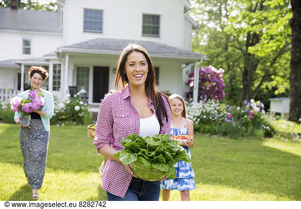 Family Party. Parents And Children Walking Across The Lawn Carrying Flowers  Fresh Picked Vegetables And Fruits. Preparing For A Party.
