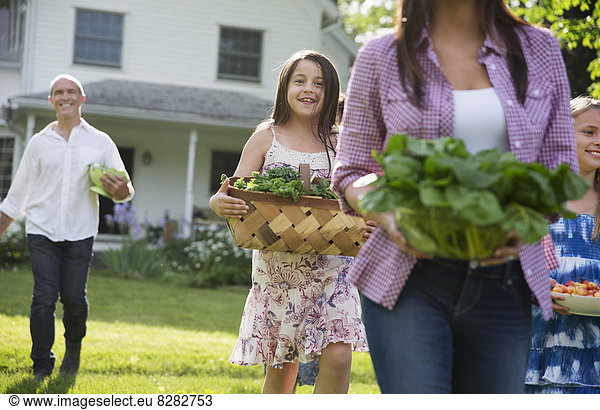 Family Party. Parents And Children Walking Across The Lawn Carrying Flowers  Fresh Picked Vegetables And Fruits. Preparing For A Party.