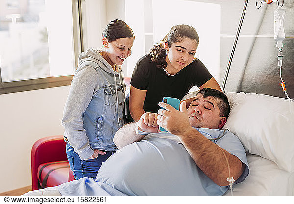 Family visiting man with cell phone lying in hospital bed