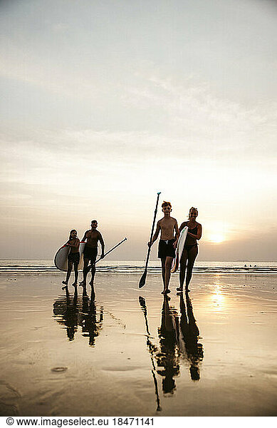 Family with paddleboards walking against sky during sunset at beach