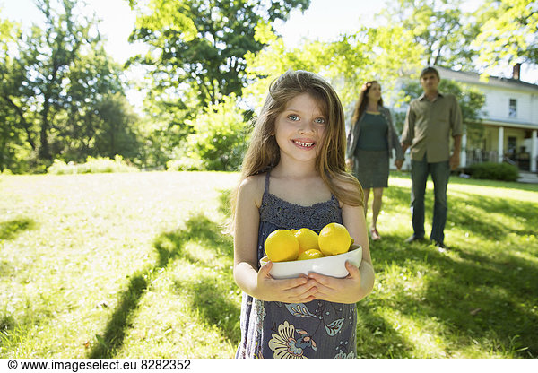 Farm. Children And Adults Working Together. A Girl Holding A Crate Of Lemons  Fresh Fruits. Two Adults In The Background.