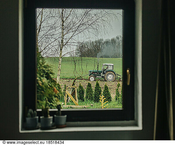 Farmer ploughing with tractor in field seen through window