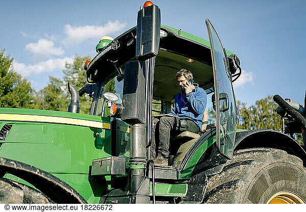Farmer using tablet PC and talking on smart phone while sitting in tractor at farm