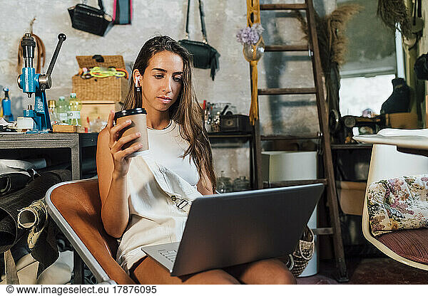 Fashion designer holding disposable coffee cup using laptop at workshop