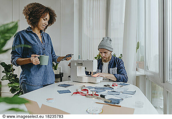 Fashion designer working on sewing machine with colleague examining denim fabric in workshop