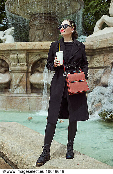 Fashion portrait of young woman standing on the wall by the fountain