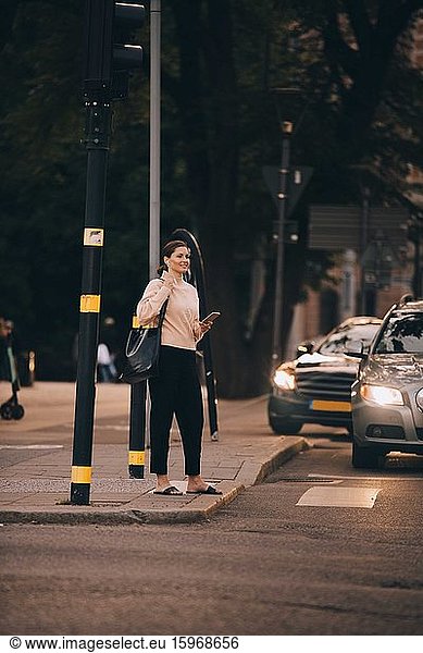 Fashionable woman with phone standing on footpath in city