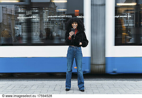 Fashionable young woman using smart phone in front of moving tram