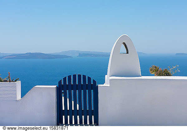 Fassade aus Backsteinzaun oder kleiner weißer Mauer mit einem blauen Tor zum Ein- oder Ausgang und einem weißen rauchfreien Schornstein mit dem blauen Ägäischen Meer im Hintergrund auf der idyllischen Insel Santorin in Griechenland