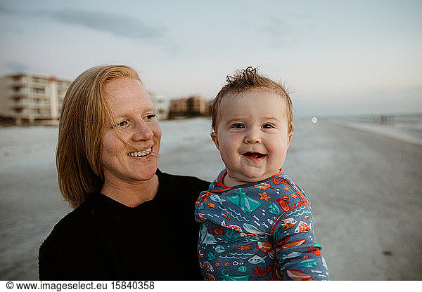 fat baby boy with crooked smile held by young red haired mom on beach