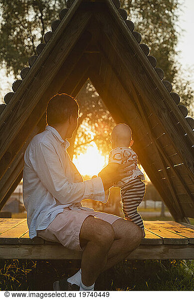 Father and baby boy sitting in wooden cabin and watching sunset at park