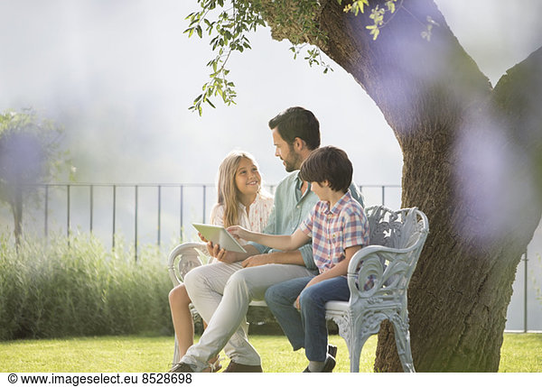 Father and children using digital tablet on bench in park