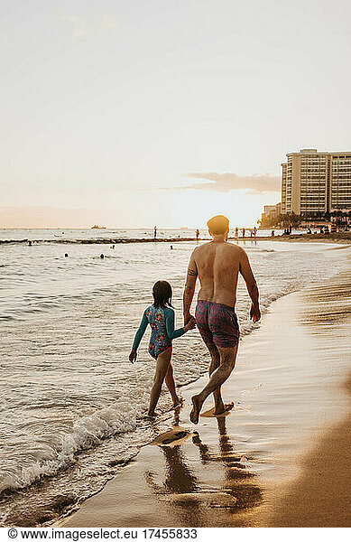 Father and daughter hold hands while they walk along beach at sunset