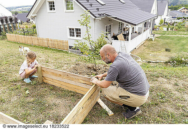 Father and daughter making raised bed crouching in back yard near house