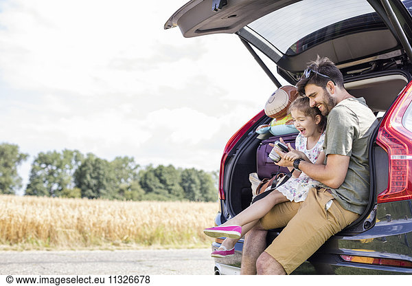 Father and daughter sitting in open car boot looking at camera at break of a road trip