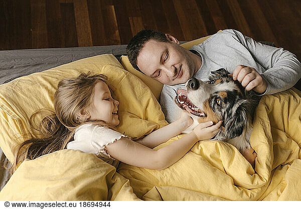 Father and daughter stroking dog lying on bed at home